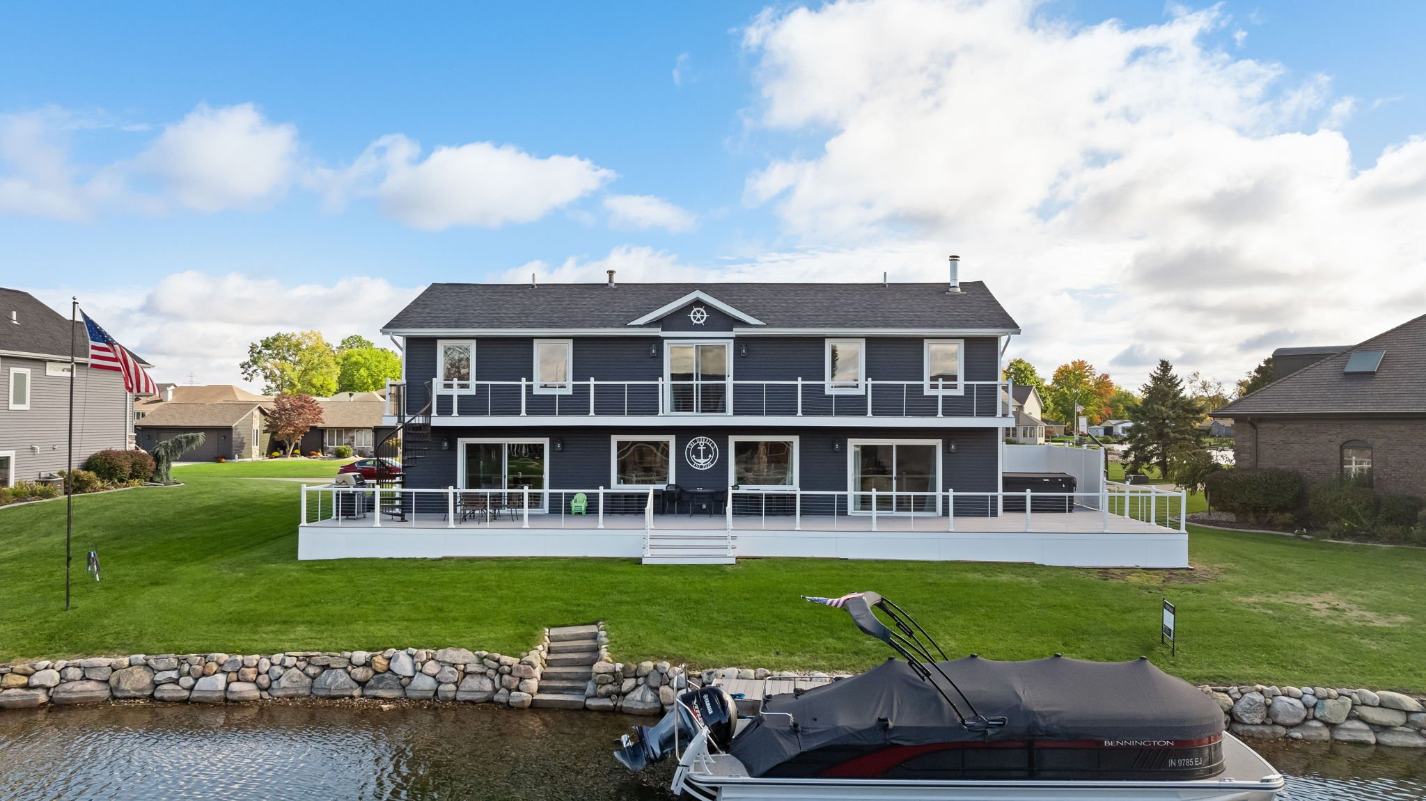 Front-facing view of a two-story waterfront home in Elkhart with a large deck, dock, and American flag by the shoreline.