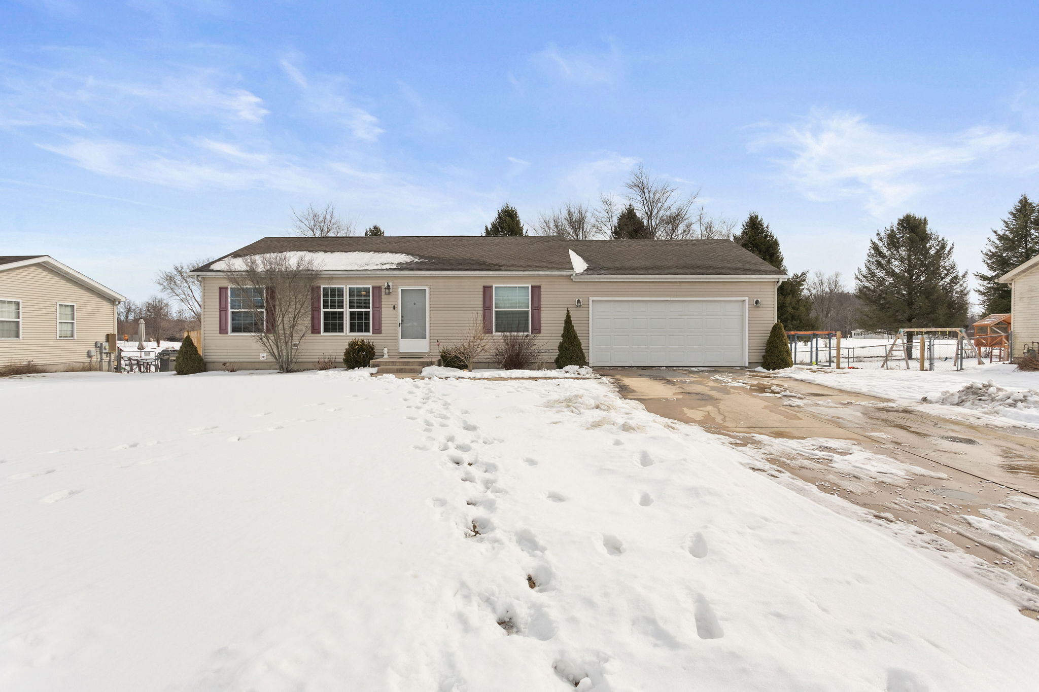 Single-story beige home with snow-covered front yard, attached two-car garage, and footprints in snow leading to front door