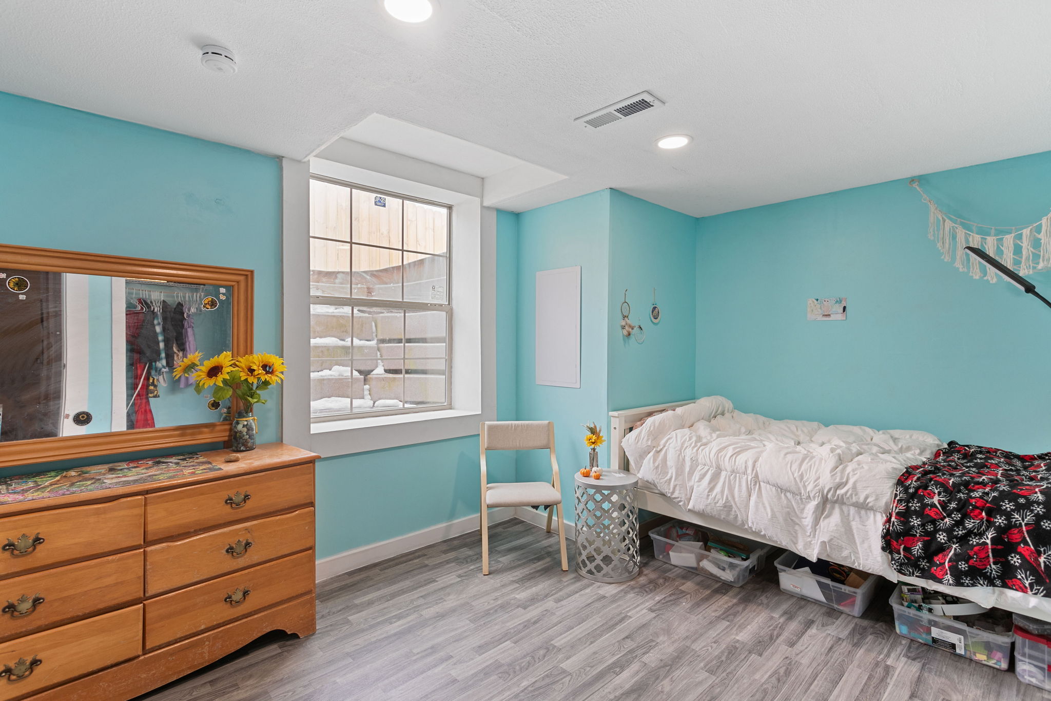 Bright basement bedroom featuring a large window, white twin bed, wooden dresser, and sunflowers in a vase.