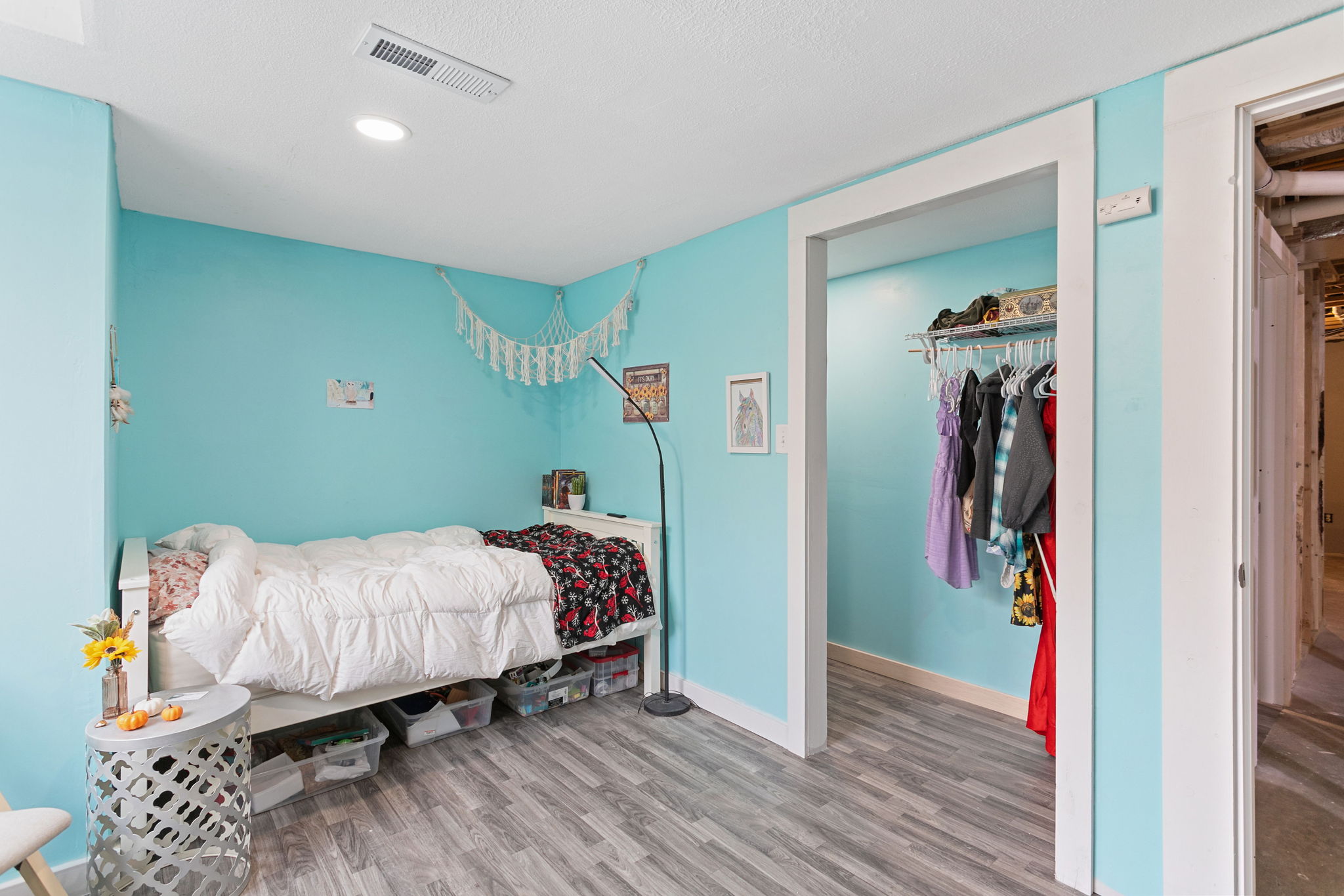 Basement bedroom with turquoise walls, white bed, and open closet filled with hanging clothes.