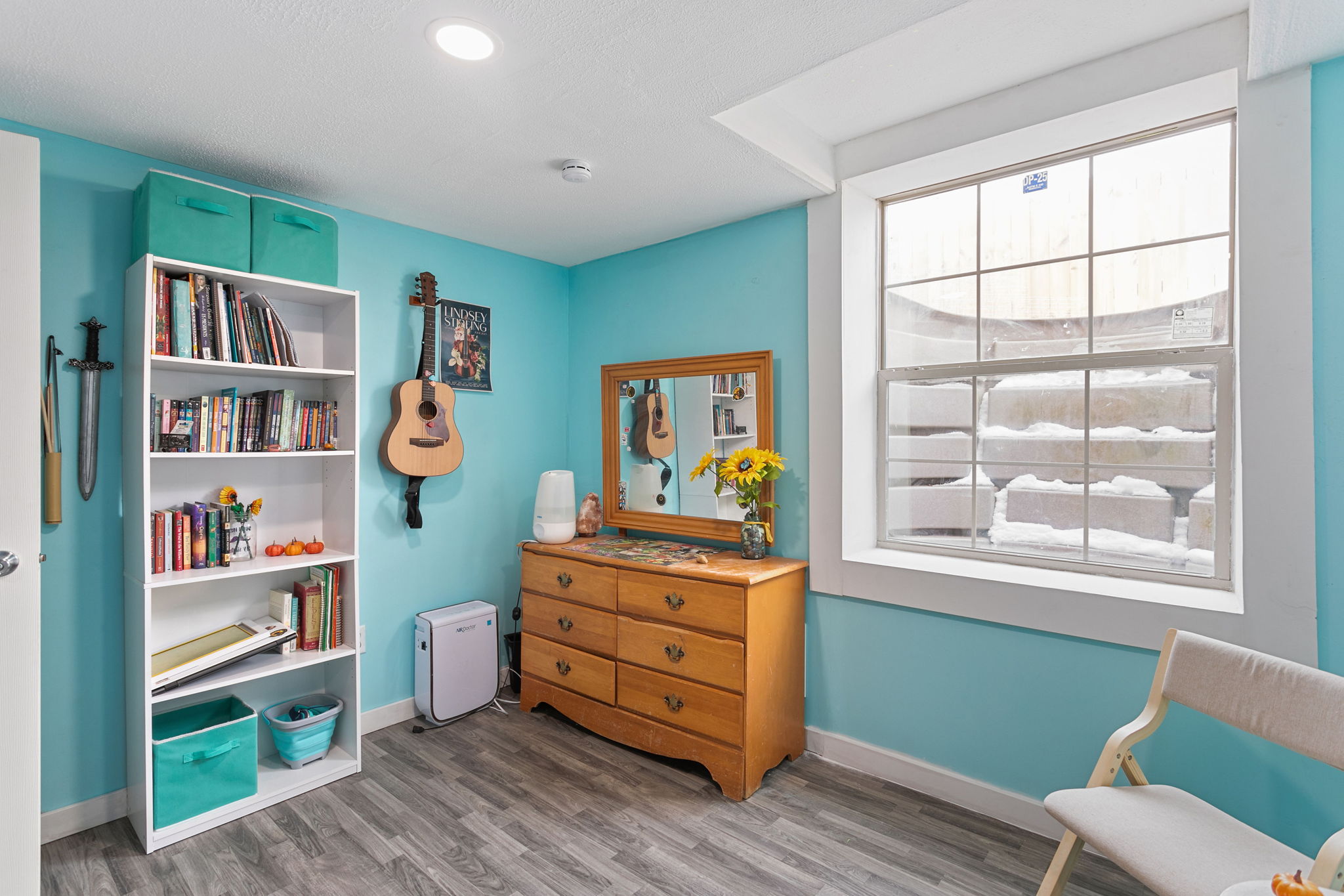 Corner view of a basement bedroom with bookshelf, acoustic guitar, and dresser beneath a window well.