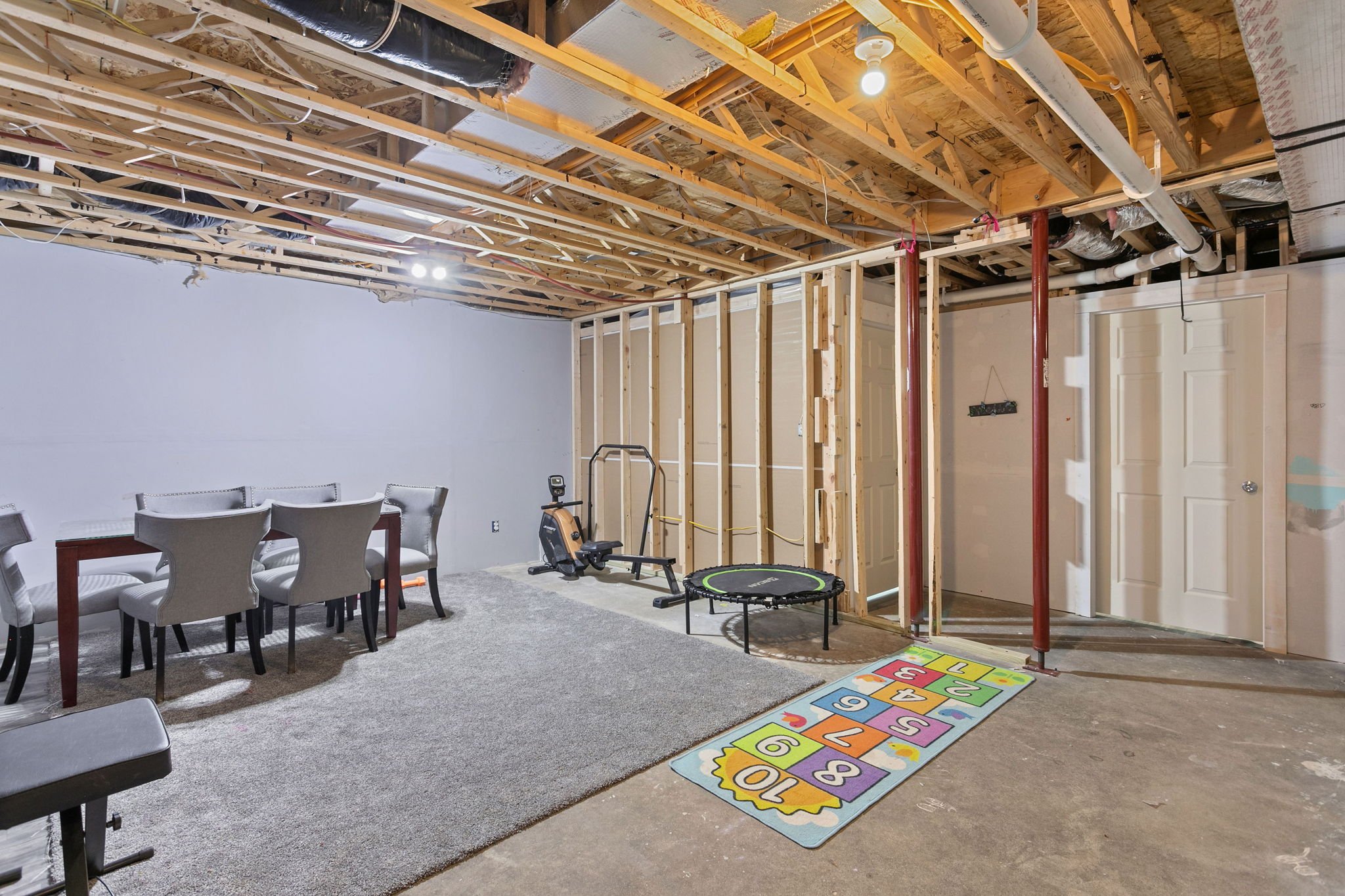 Unfinished basement area with dining table, home gym equipment, trampoline, and open framing.