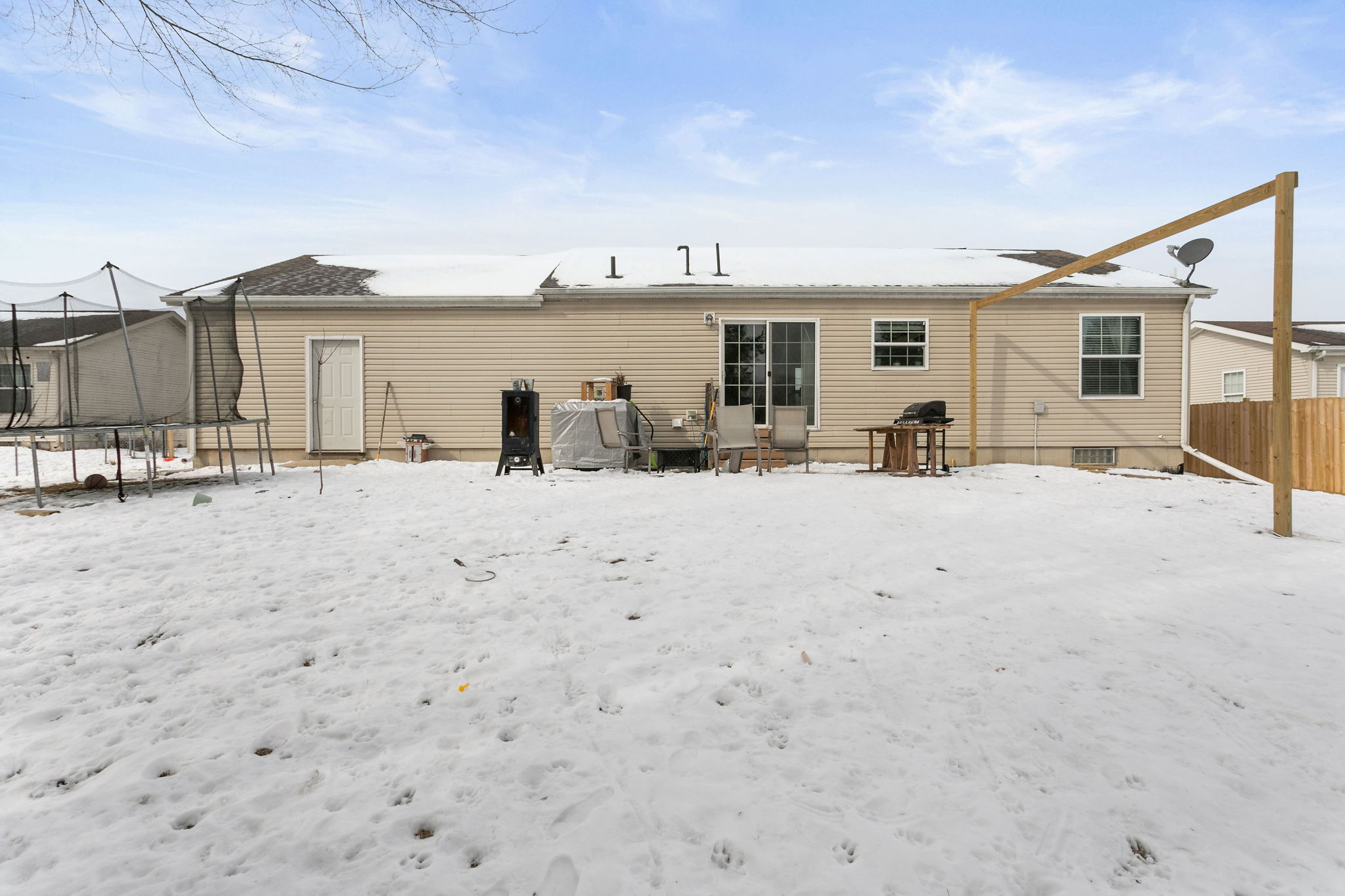 Backyard view of beige single-story home in winter, with snow-covered lawn and patio furniture.