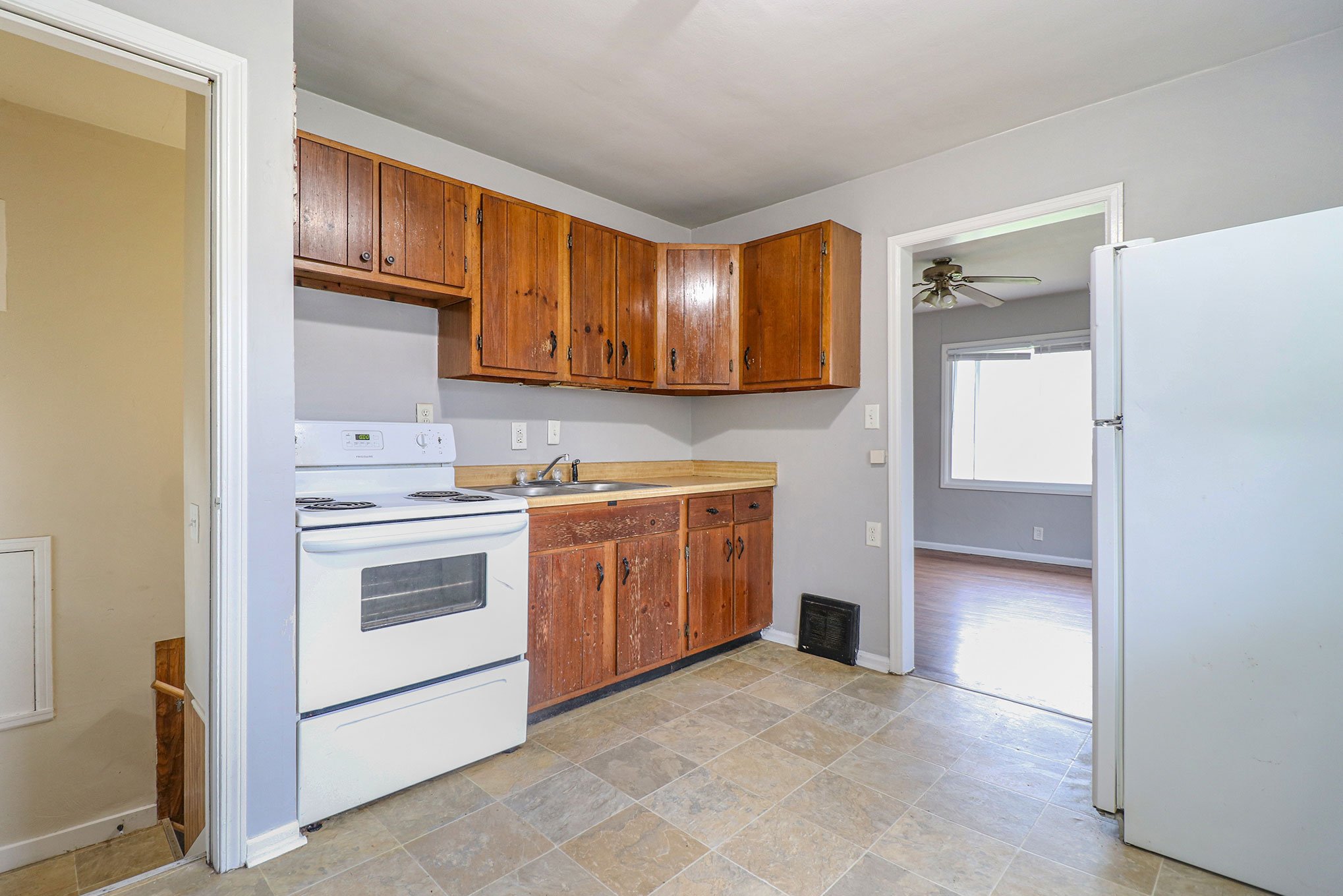 Modest kitchen with wood cabinets, white appliances, and tile flooring, adjacent to a stairwell.
